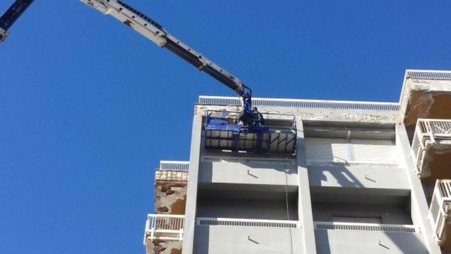 Una gru solleva una piattaforma accanto a un edificio, probabilmente per lavori di costruzione o riparazione. Sullo sfondo il cielo azzurro.