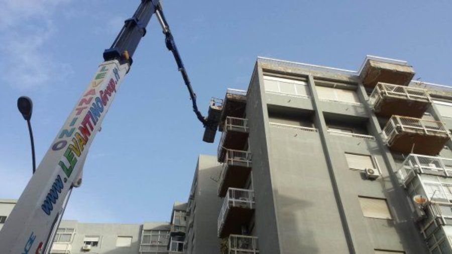 Una gru solleva una piattaforma fino al balcone di un condominio grigio in una giornata di sole.
