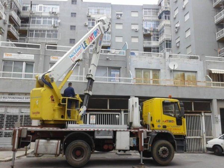 Autogru gialla con operatore, parcheggiata in strada. Edifici sullo sfondo.