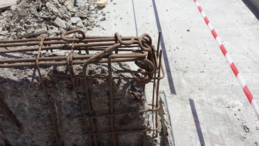 Armatura di rinforzo esposta nell'angolo di una struttura in cemento con resti di cemento, accanto a una barriera a strisce bianche e rosse.