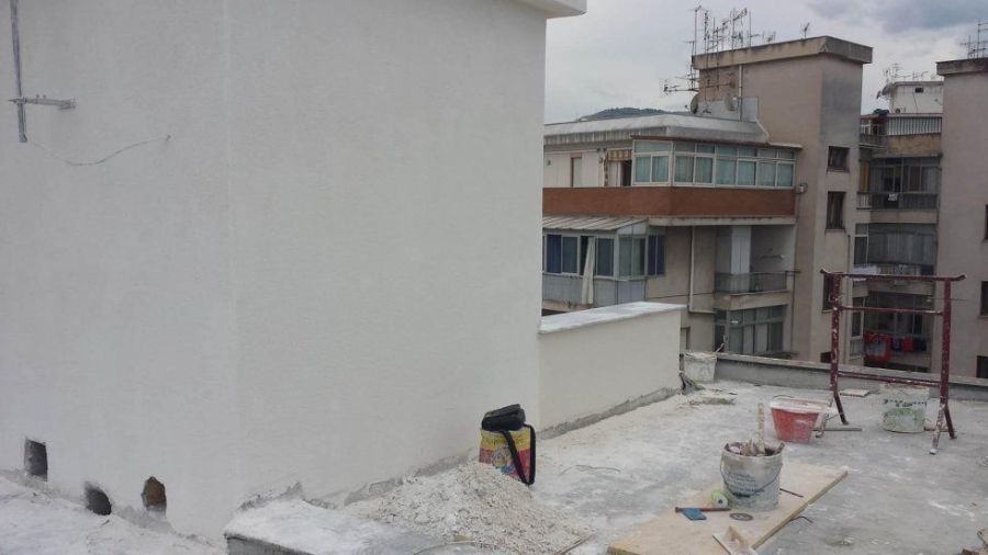 Vista dal tetto di diversi edifici bianchi sotto un cielo coperto. Secchi di vernice e materiali sparsi sul tetto.