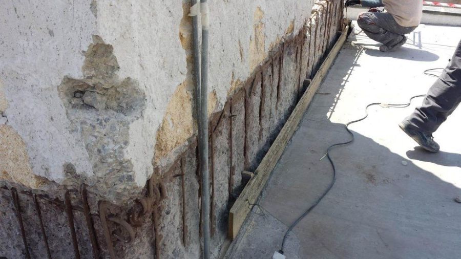 Armatura di rinforzo esposta in un muro di cemento danneggiato, con una persona che lavora nelle vicinanze e una cassaforma di legno.