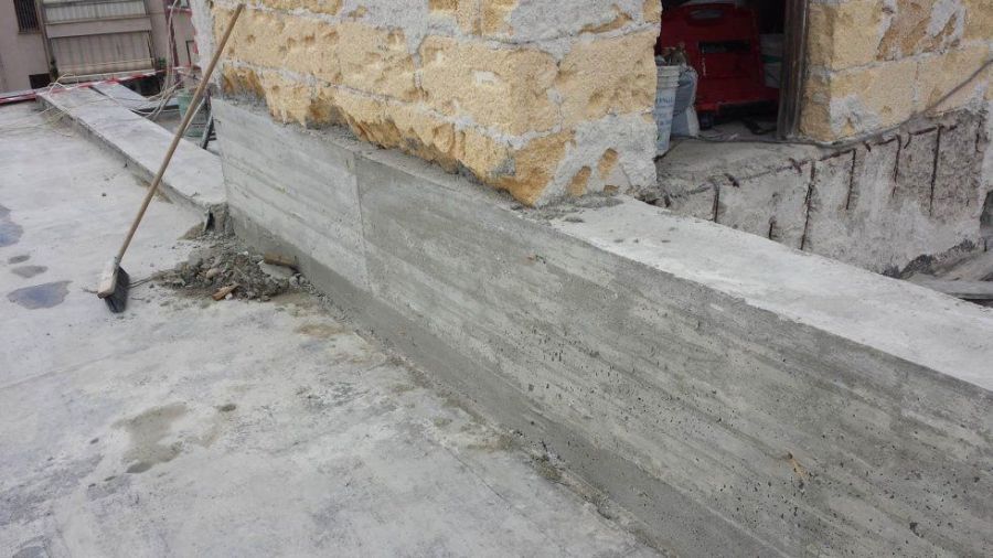 Un muro di contenimento in cemento armato in costruzione accanto a un edificio in pietra usurato. Sono presenti materiali da costruzione e attrezzi.