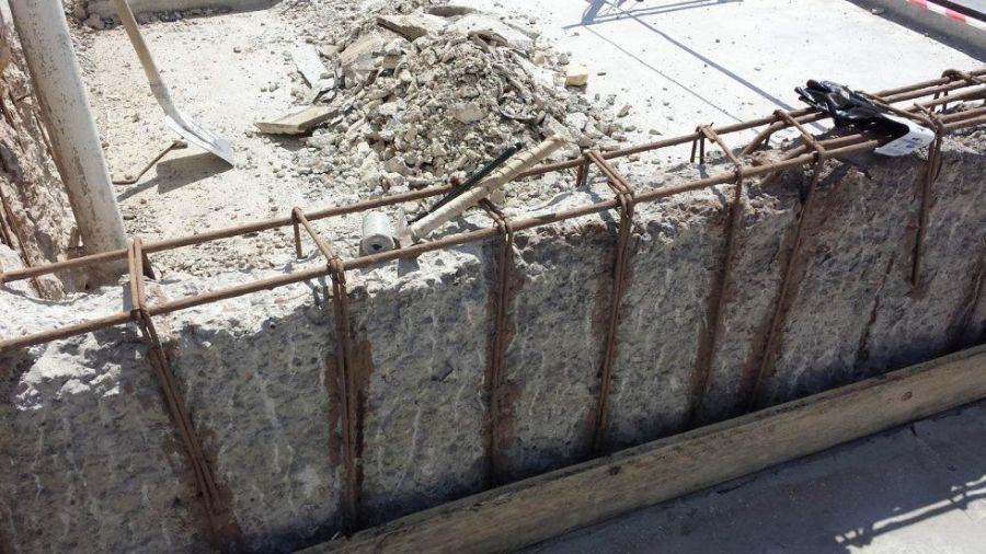 Cantiere con barre di armatura esposte in un muro di cemento. Sono presenti detriti e attrezzi.