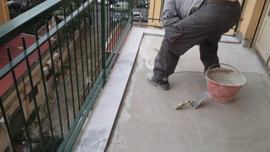 Persona che stende il cemento sul pavimento di un balcone. Il balcone ha una ringhiera verde e degli edifici visibili. Sono presenti un secchio e una cazzuola.