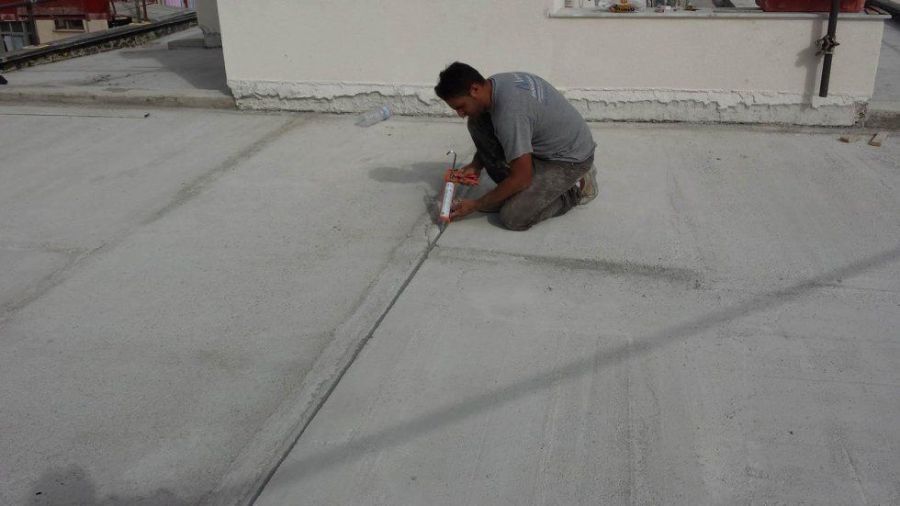 Uomo inginocchiato su una superficie di cemento, mentre applica del sigillante lungo una giunzione. Sullo sfondo un edificio bianco.