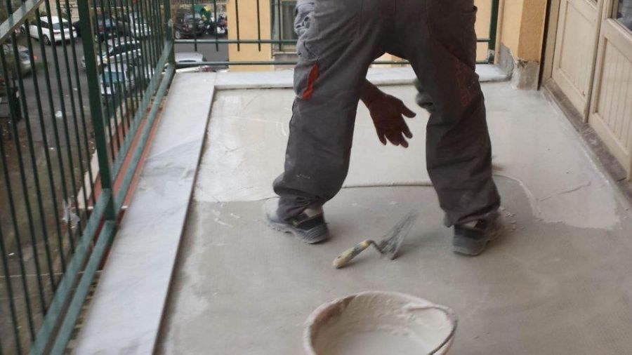 Persona che stende il cemento sul pavimento di un balcone con una cazzuola; sono visibili il secchio e la ringhiera.