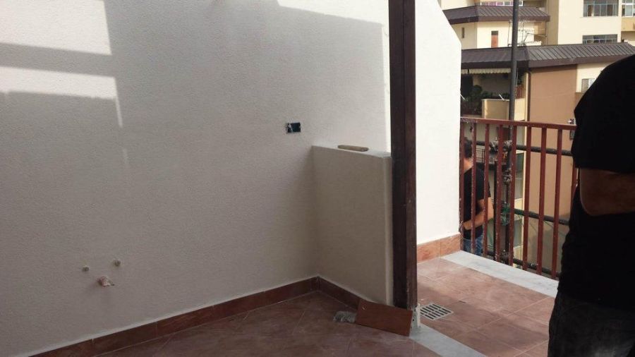 Angolo vuoto di una stanza con pareti bianche, pavimento in piastrelle marroni e una porta che conduce a un balcone.