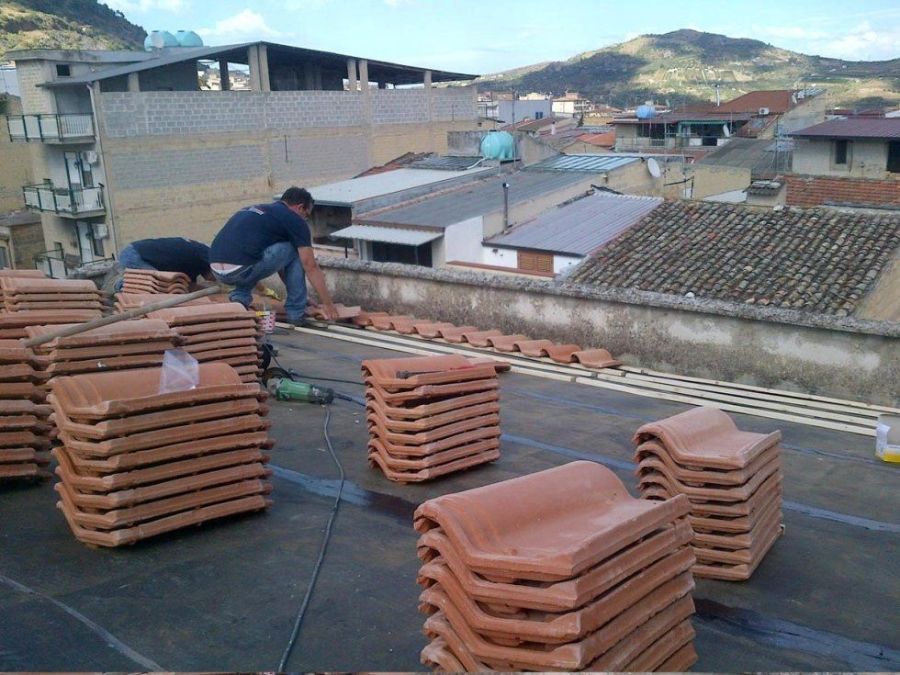 Operai che installano tegole in terracotta su un tetto piano, circondato da pile di tegole, in un contesto urbano.