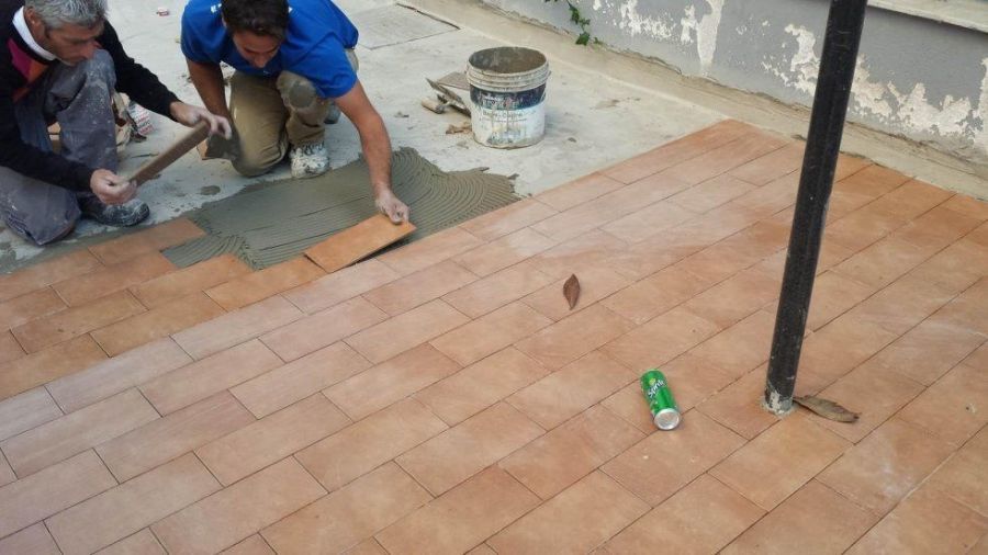 Due operai posano piastrelle color terracotta su una superficie di cemento all'esterno; uno è inginocchiato e stende la malta, l'altro usa un martello.