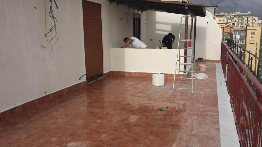 Lavoratori su una terrazza parzialmente ristrutturata; pavimento piastrellato rosso, pareti bianche, porta marrone e vista sulla città.