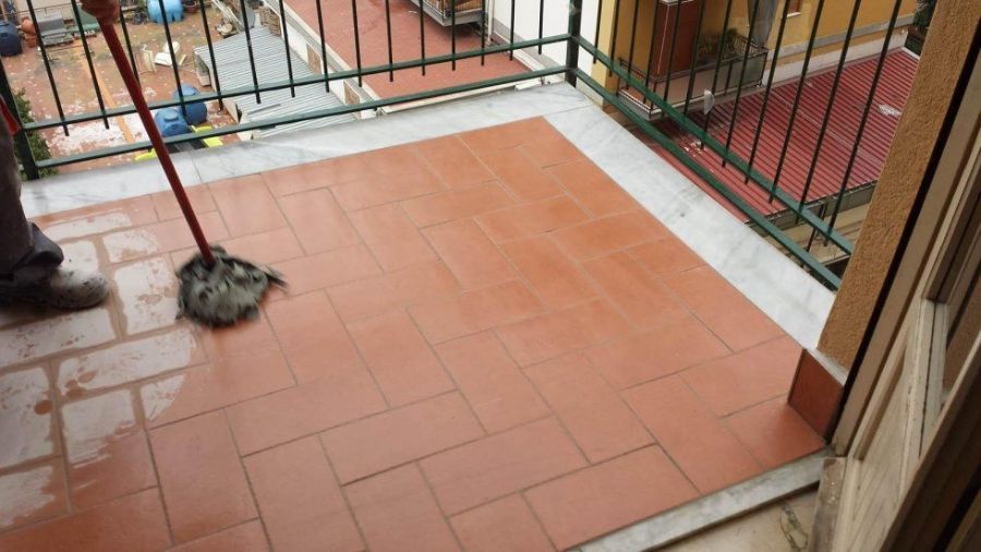 Persona che lava un balcone piastrellato. Sono visibili il pavimento bagnato e la ringhiera metallica.
