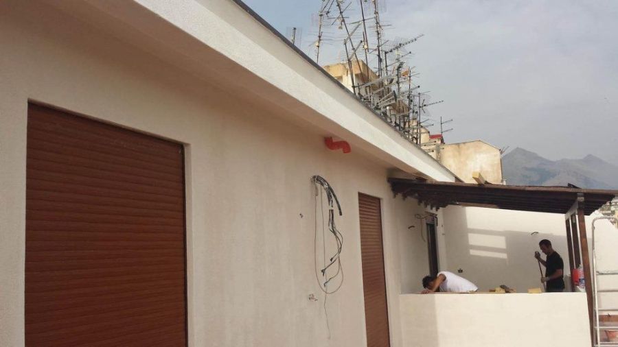 Edificio marrone chiaro con una persiana marrone, una porta aperta e due uomini, uno appoggiato e uno in piedi fuori.