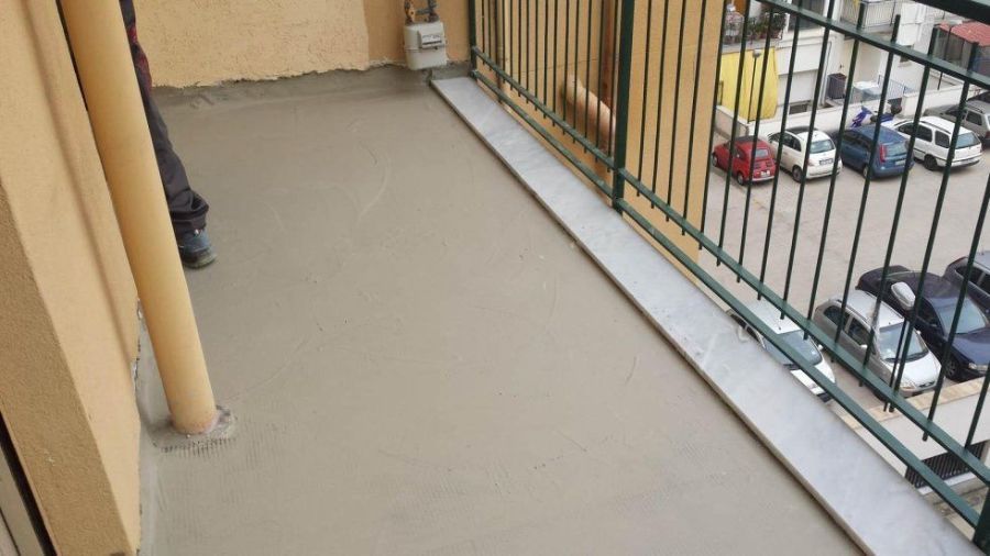 Balcone con pavimento in cemento grigio chiaro e ringhiera in metallo che si affaccia sulle auto parcheggiate.