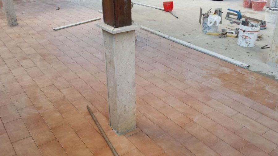 Area esterna piastrellata in costruzione; piastrelle marroni, pilastro di cemento, attrezzi e secchi visibili.