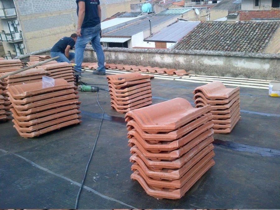 Pile di tegole rosse su un tetto piano; due operai le stanno installando.