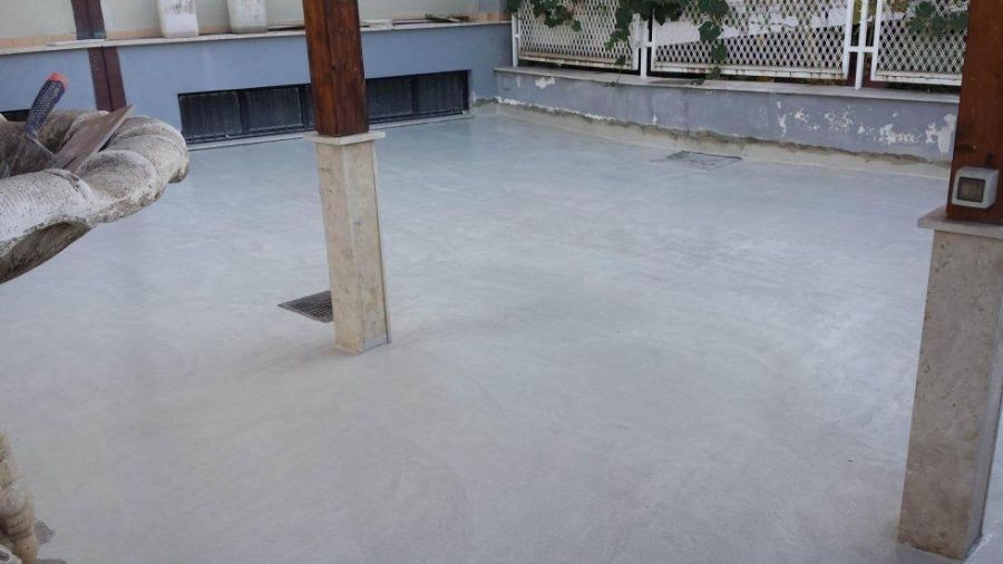 Un pavimento in cemento grigio chiaro appena gettato in uno spazio esterno con pilastri di sostegno e un muro.