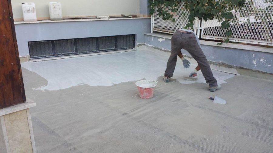 Persona che applica una mano di vernice bianca su una superficie di cemento all'aperto. Sono visibili secchio, attrezzi e un muretto.