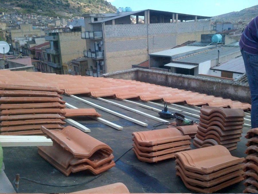 Tetto con pile di tegole di argilla arancione, sbarre di metallo e una persona che lavora; edifici e collina sullo sfondo.