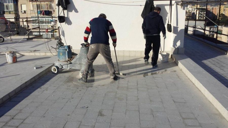 Due persone su un tetto applicano del materiale con degli attrezzi sulla superficie piastrellata, accanto a un serbatoio di gas e a un muro.