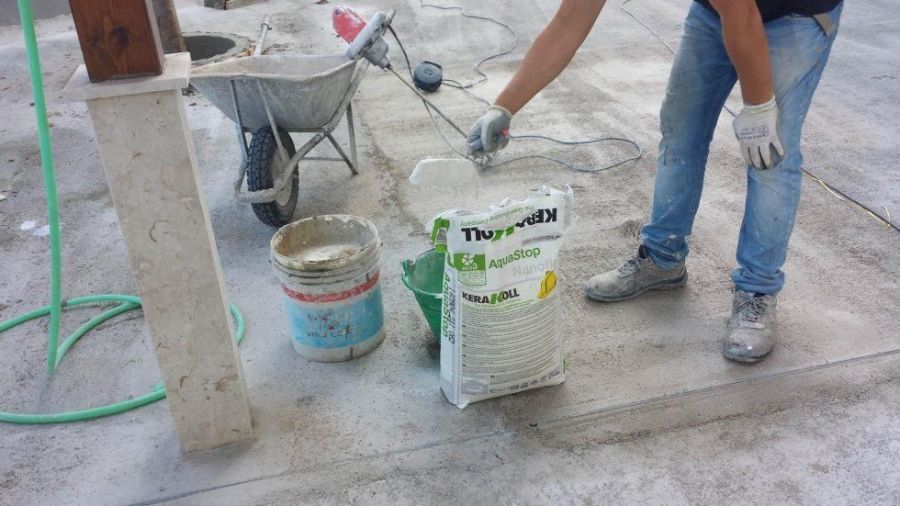 Persona in jeans e guanti che lavora con il cemento vicino a una carriola e a dei sacchi su una superficie di cemento.