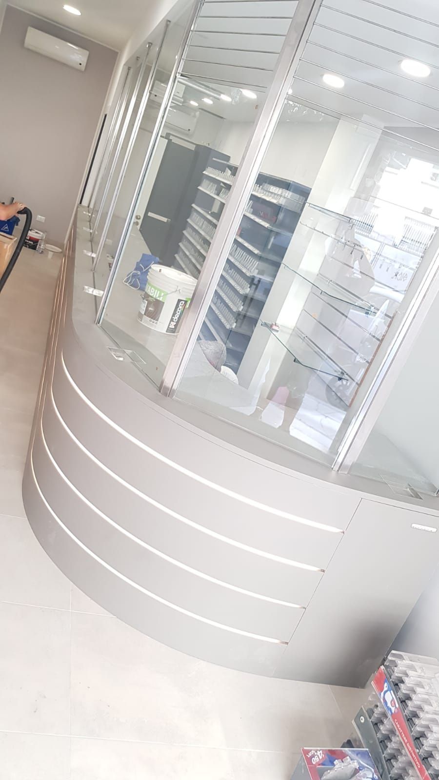 Bancone da farmacia curvo e grigio con vetrine; scaffali con prodotti visibili all'interno.