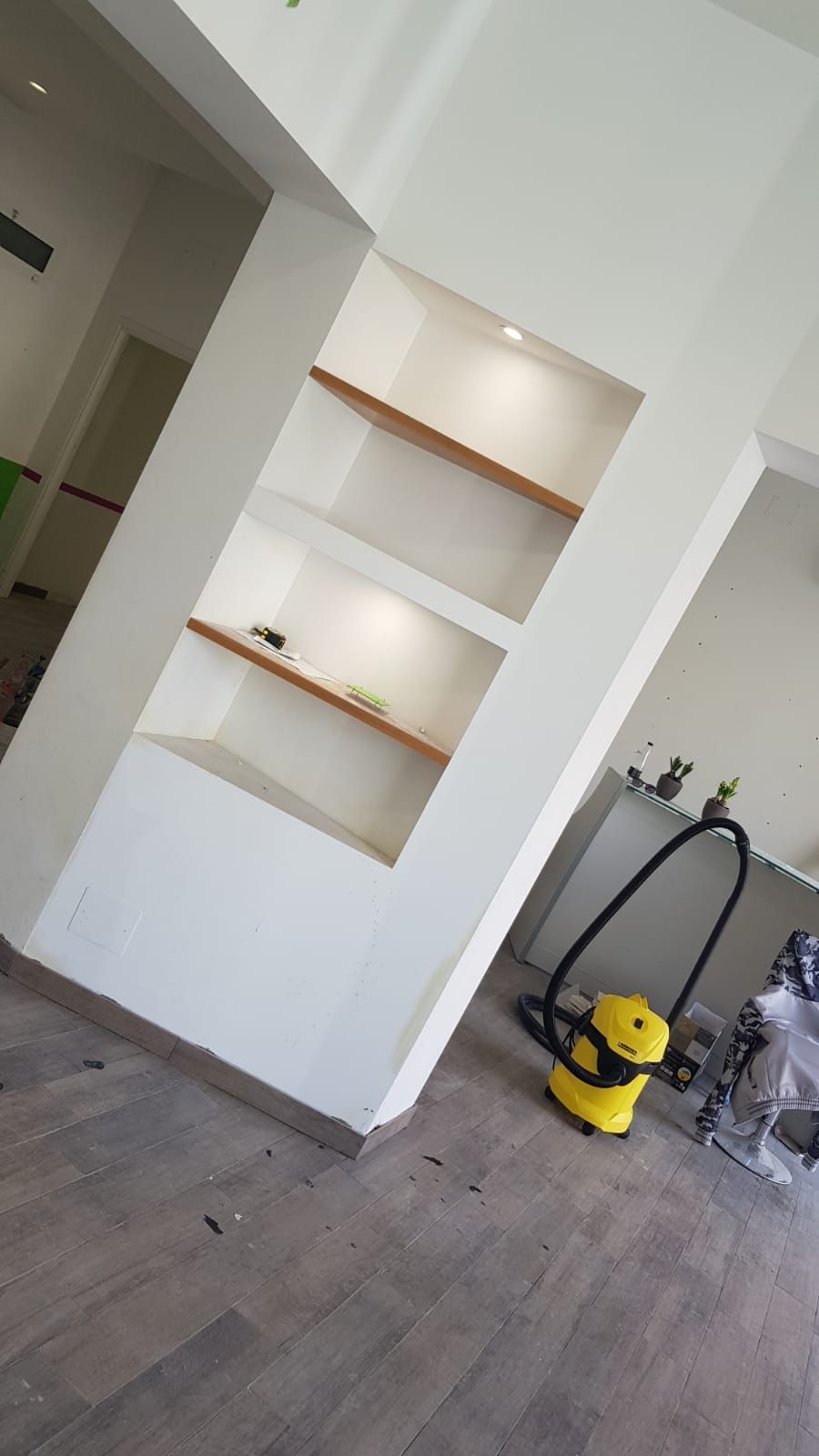 Mensole bianche a muro con ripiani in legno su una parete bianca. Un aspirapolvere giallo è appoggiato lì vicino.