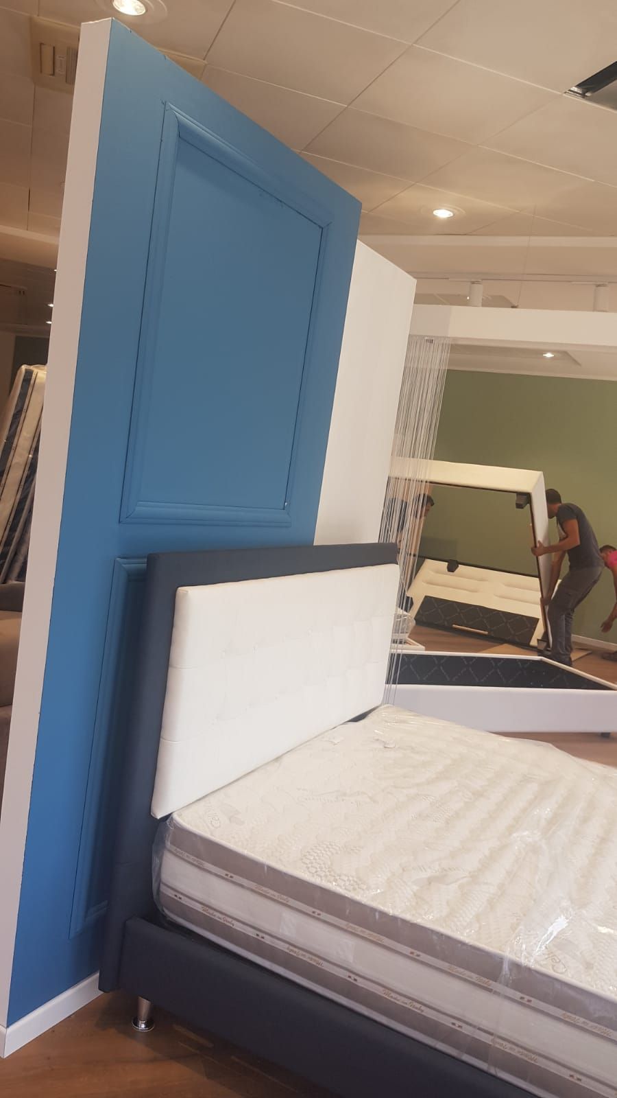 Letto con materasso bianco e testiera blu in un negozio di mobili. Persone sullo sfondo.