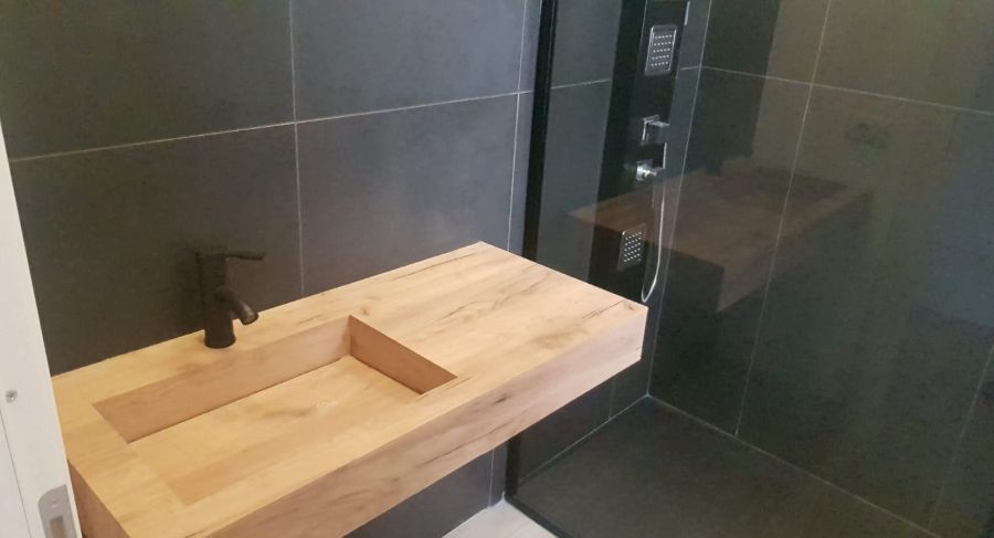Bagno con lavandino in legno, rubinetto nero e doccia dietro un pannello di vetro su una parete piastrellata nera.