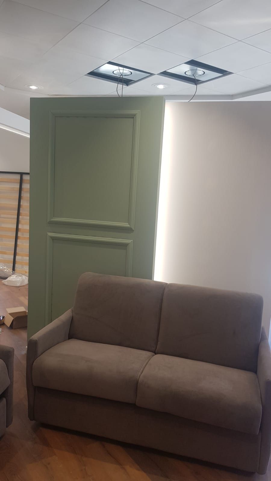 Porta verde, divano marrone contro una parete bianca. Luci a soffitto.