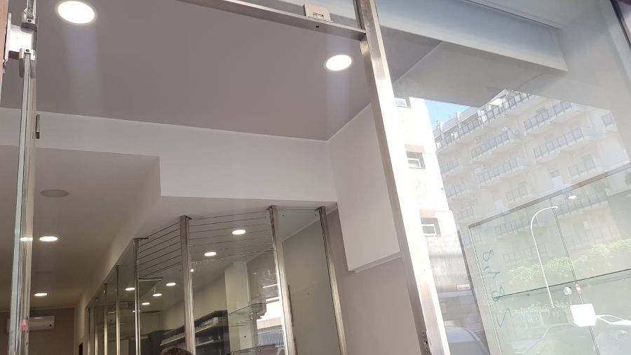 Vista interna del negozio con porte in vetro, soffitto bianco e scorcio di un edificio esterno.