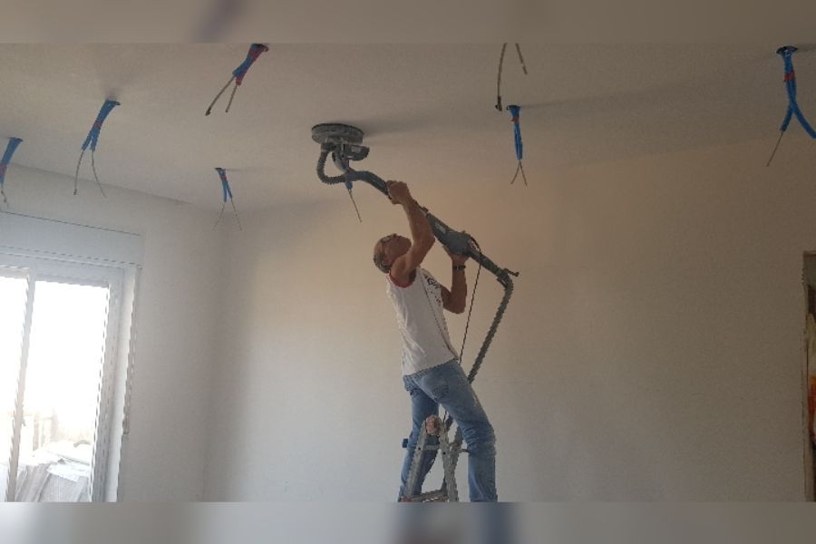 Un uomo sta levigando un soffitto su una scala, utilizzando una levigatrice a manico lungo, in una stanza con fili elettrici scoperti.