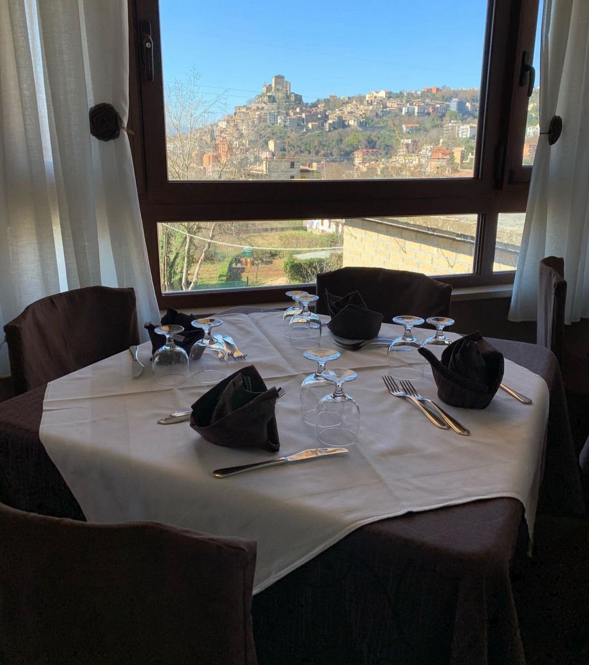 Tavolo apparecchiato per cenare con vista su una cittadina collinare attraverso una finestra.