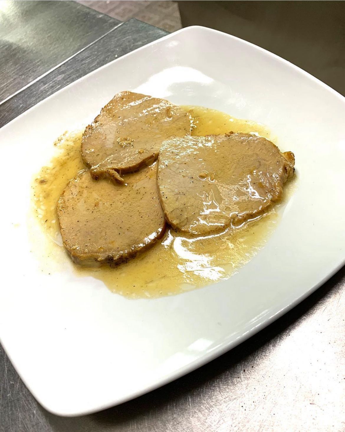 Tre fette ovali di carne immerse in una salsa chiara su un piatto rettangolare bianco.