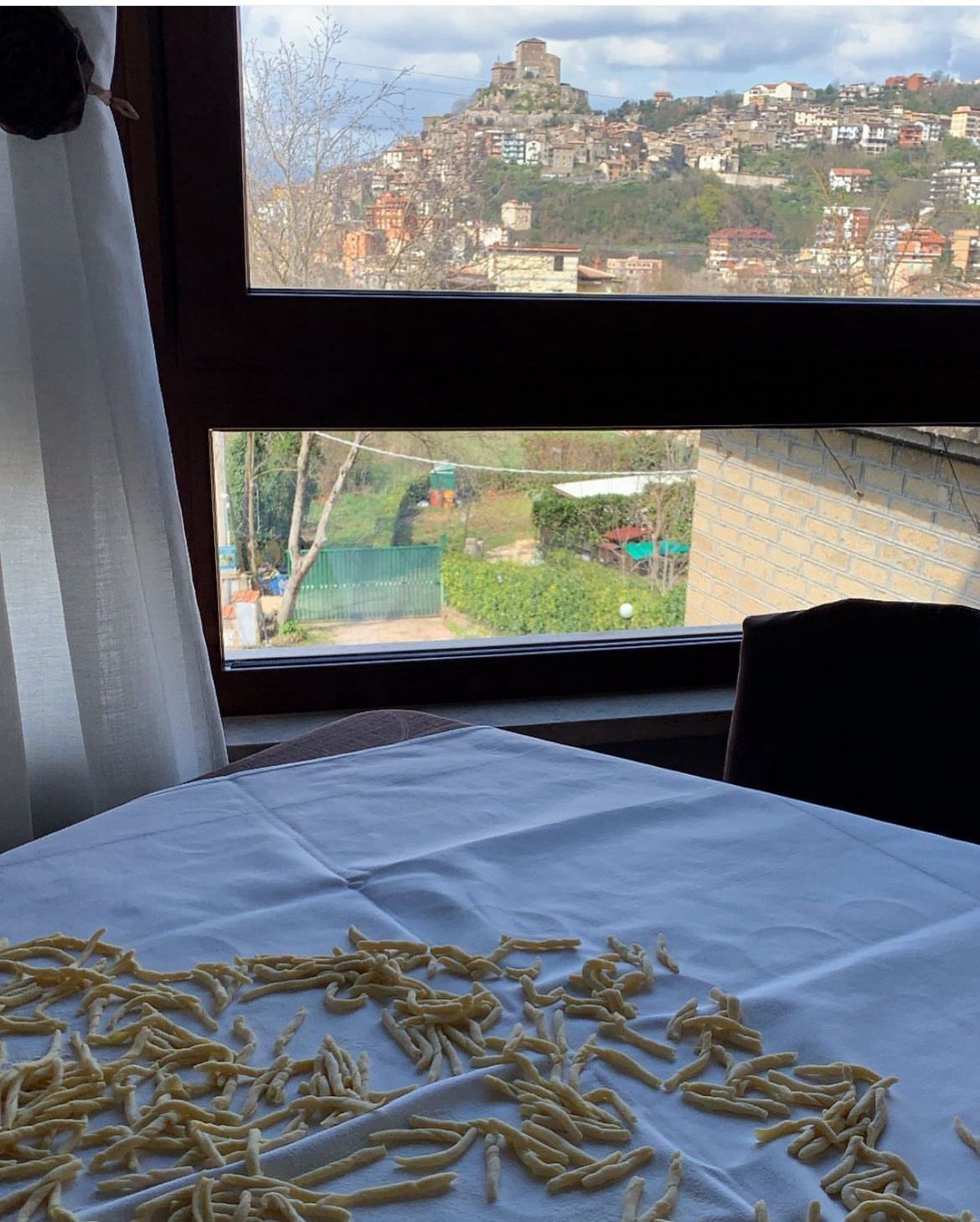 Pasta su una tovaglia bianca con vista dalla finestra su una cittadina in cima a una collina e cielo azzurro.