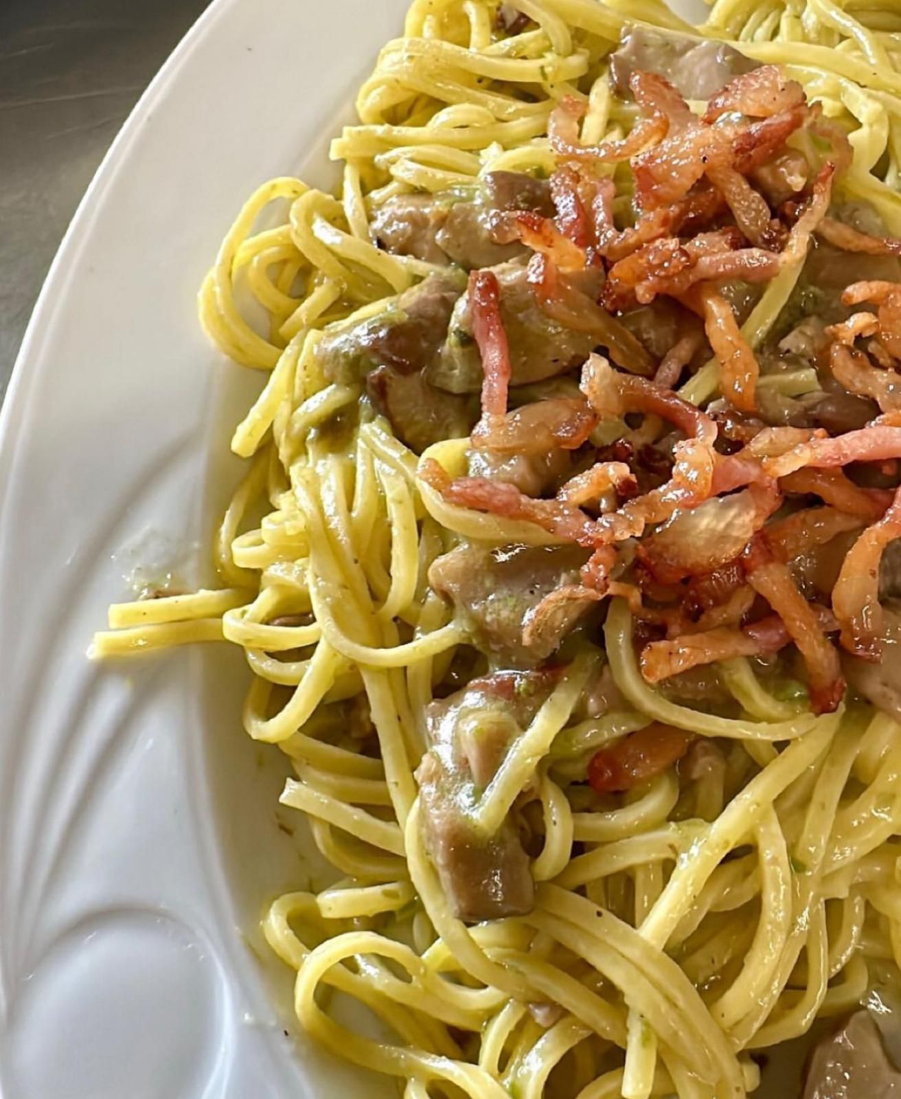 Piatto di pasta con salsa cremosa, funghi e pancetta croccante su un piatto bianco.