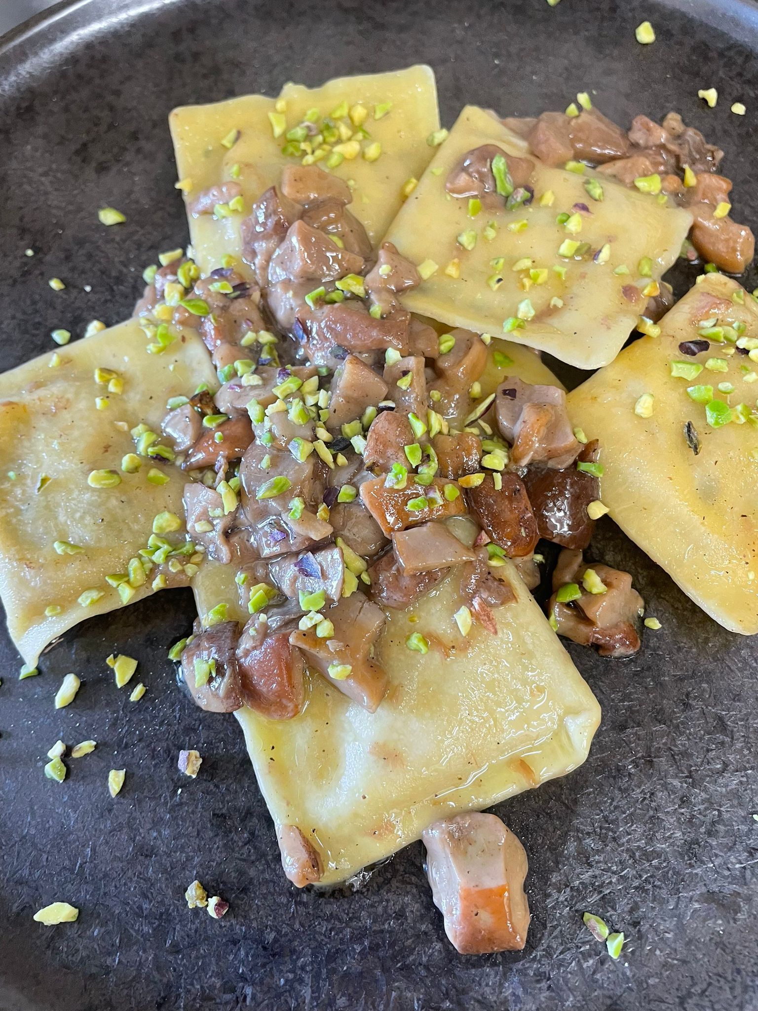 Ravioli con salsa ai funghi e crumble di pistacchi, serviti su un piatto scuro.