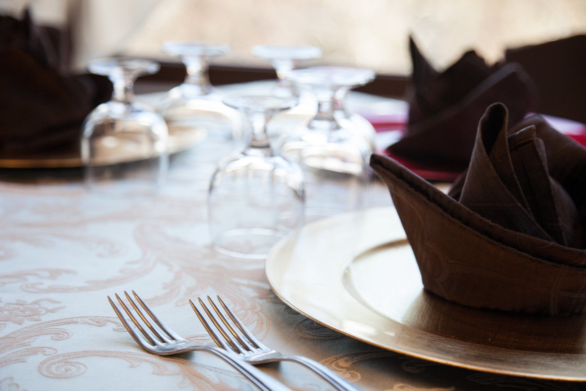 Tavola apparecchiata per un pasto con piatto dorato, tovagliolo marrone, bicchieri e forchette d'argento.