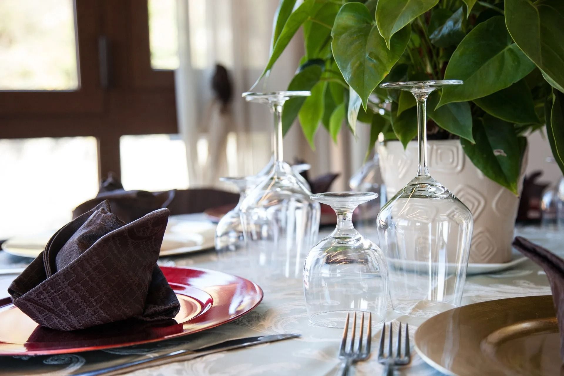 Un tavolo apparecchiato per cenare con piatti rossi e oro, tovaglioli piegati e bicchieri da vino, accanto a una pianta in vaso.