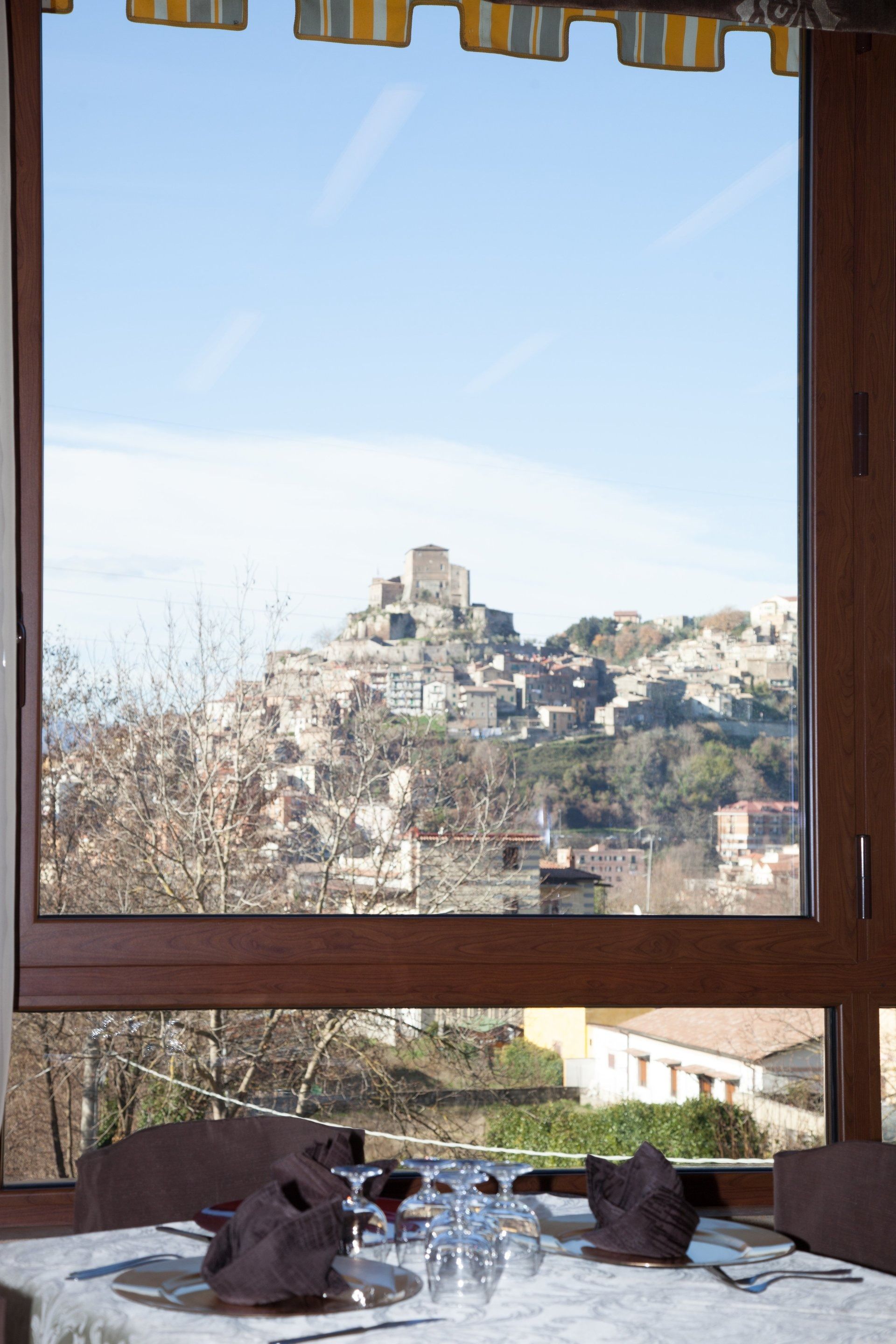 Vista da una finestra di un tavolo apparecchiato per cenare, con un villaggio storico in cima a una collina visibile in lontananza.