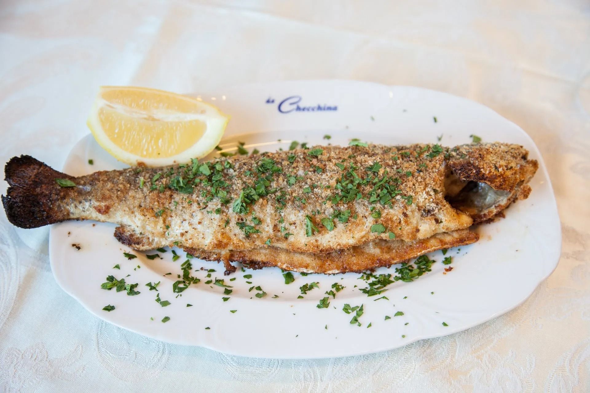 Pesce intero al forno con spicchio di limone su un piatto bianco, cosparso di prezzemolo.