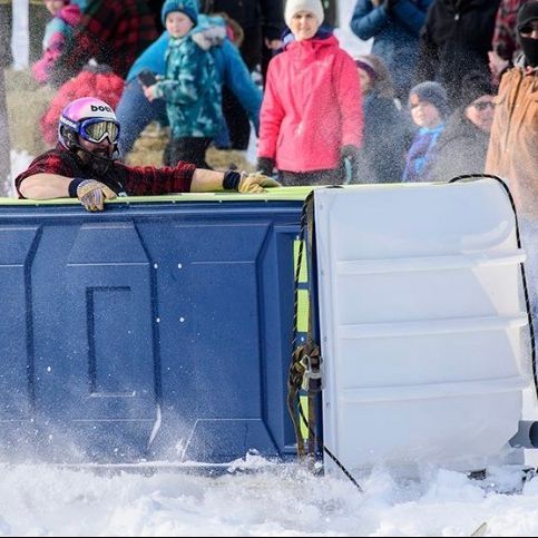 Man in a portable toilet racing across snow, wearing goggles. Spectators watch in the background.
