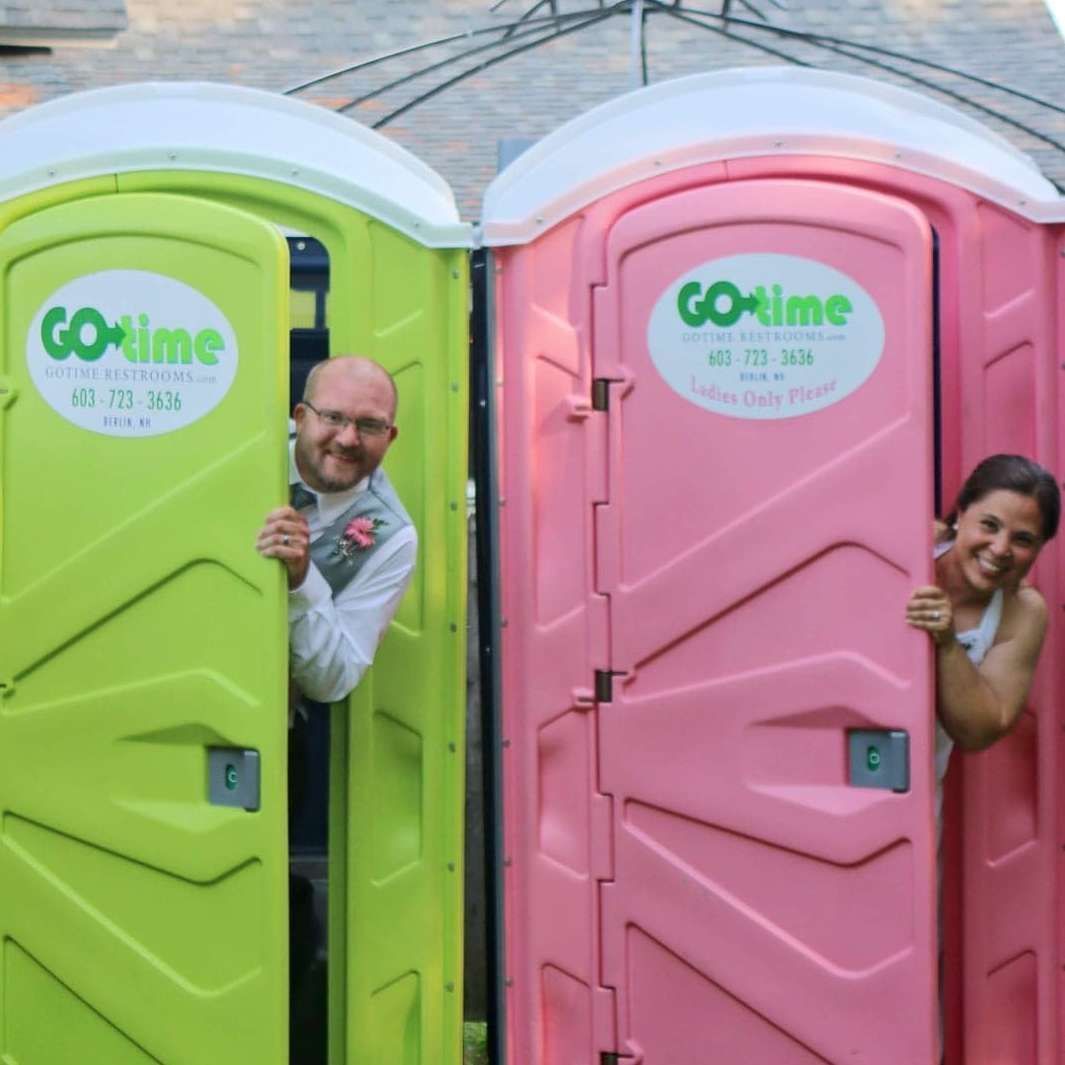 A newlywed couple peeks out from green and pink portable toilets at an outdoor wedding, smiling.