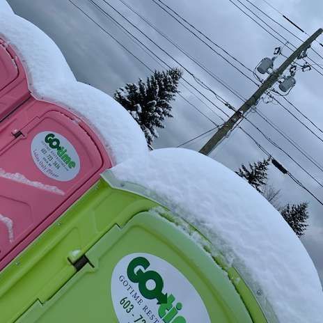 Two colorful portable toilets, pink and green, covered in snow. Power lines and trees in background.