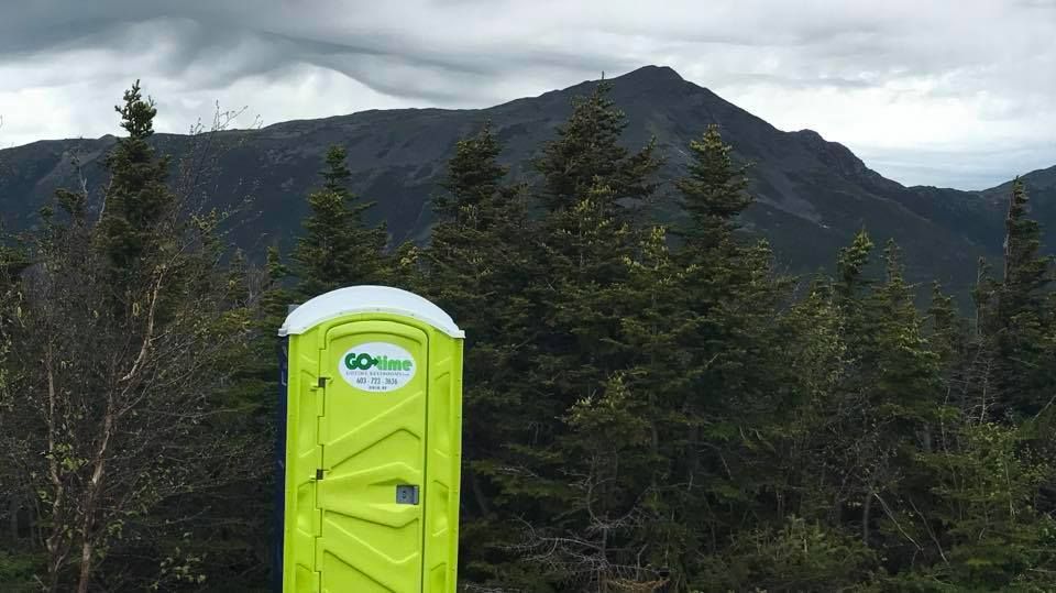 Lime green portable toilet in a mountain forest with a peak in the background under a cloudy sky.