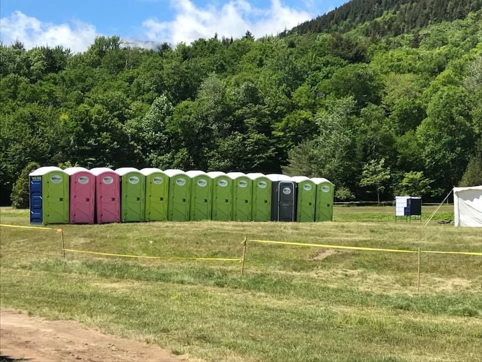 Row of colorful portable toilets on a grassy field with a backdrop of trees and mountains.