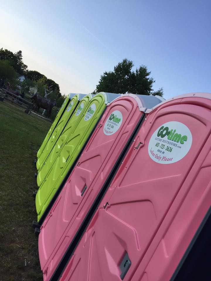Row of colorful portable toilets: lime green, pink, and white, on a lawn.