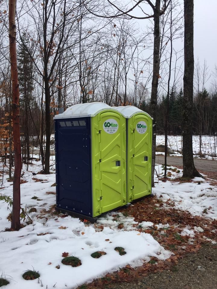 Two lime green and blue portable toilets sit in a snowy, wooded area.