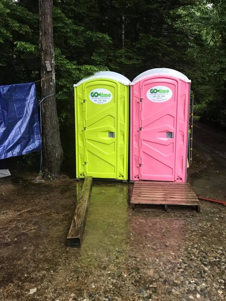 Two portable toilets, one green, one pink, sit outdoors near a tree and tarp on a rainy day.
