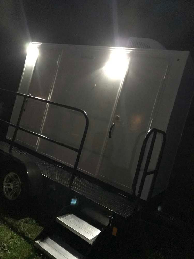 Portable restroom trailer at night with illuminated entry steps and exterior lights.